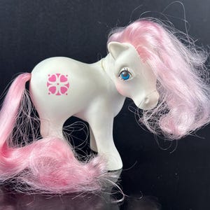 May include: A white toy pony with pink hair and tail. The pony has blue eyes and a pink floral design on its side. The mane and tail are long and flowing. The toy is a vintage collectible.