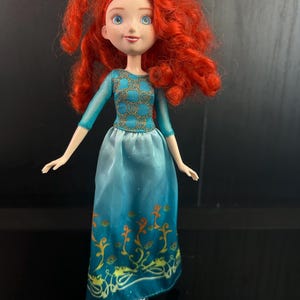 May include: A doll with bright red curly hair, blue eyes, and freckles. The doll wears a teal dress with gold accents on the bodice and a decorative pattern on the skirt. The doll is standing on a clear plastic stand.