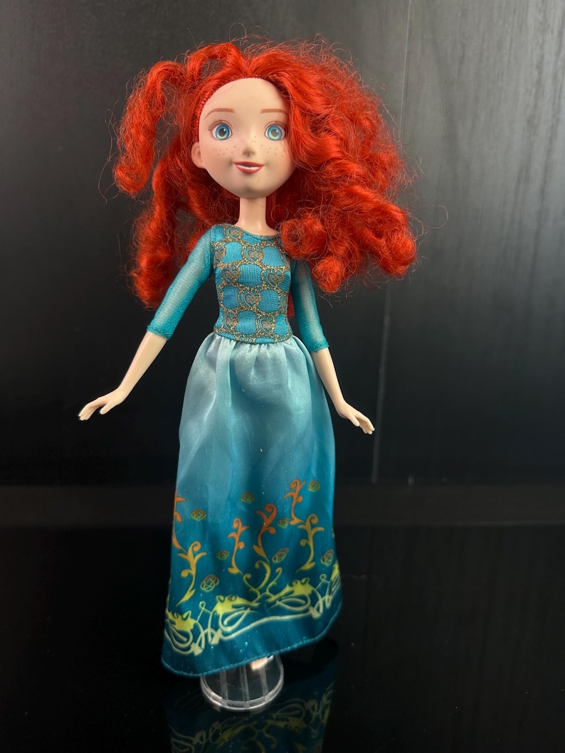 May include: A doll with bright red curly hair, blue eyes, and freckles. The doll wears a teal dress with gold accents on the bodice and a decorative pattern on the skirt. The doll is standing on a clear plastic stand.