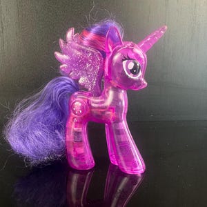 Op de afbeelding: Een doorschijnend roze speelgoedpony met paars haar en een hoorn. De pony heeft glinsterende vleugels en een lange, vloeiende staart. Het speelgoed staat op een reflecterend oppervlak.