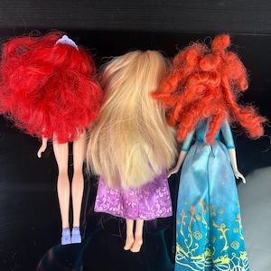 May include: Three dolls with long hair and dresses are displayed. The doll on the left has bright red hair and a purple dress. The middle doll has long blonde hair and a purple dress. The doll on the right has red curly hair and a blue dress.
