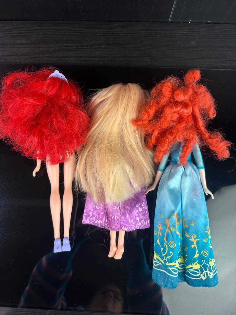 May include: Three dolls with long hair and dresses are displayed. The doll on the left has bright red hair and a purple dress. The middle doll has long blonde hair and a purple dress. The doll on the right has red curly hair and a blue dress.