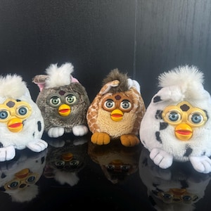 Op de afbeelding: Vier pluche Furby-speelgoed in verschillende kleuren worden getoond tegen een zwarte achtergrond. De Furby's hebben grote ogen, gele en oranje snavels en plukjes haar op hun hoofd. Eén is wit met zwarte vlekken.
