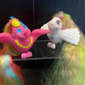 Op de afbeelding: Twee vogelbeeldjes met lang, vloeiend, meerkleurig synthetisch haar. De ene vogel is felroze met een gele ketting en een gele kuif, terwijl de andere wit is met een blauwe strik op de borst. Beide vogels hebben grote, expressieve ogen.