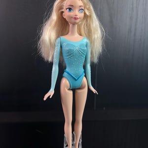 May include: A doll with long, blonde hair and blue eyes. The doll is wearing a light blue leotard with long sleeves and a decorative pattern. The doll is standing on a clear plastic stand.