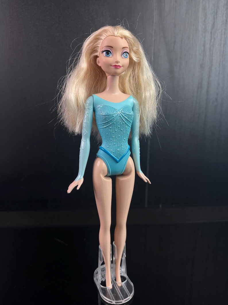 May include: A doll with long, blonde hair and blue eyes. The doll is wearing a light blue leotard with long sleeves and a decorative pattern. The doll is standing on a clear plastic stand.
