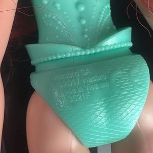 May include: Close-up of a turquoise doll's torso and legs. The doll's outfit includes a detailed bodice and a skirt-like bottom. The text "HASBRO SA &copy;2017 Hasbro Made in Vietnam" is printed on the doll's bottom.