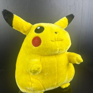 May include: A plush Pikachu toy in a sitting position. The toy is primarily yellow with black-tipped ears, a red cheek, and a smiling face. The toy is a popular character from the Pokémon franchise.