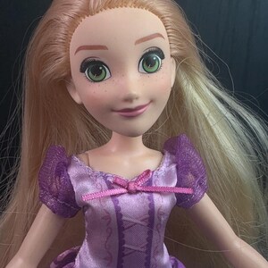 May include: A doll with long blonde hair and green eyes. The doll is wearing a purple dress with a pink bow. The doll has a smiling expression and freckles on its face. The doll's dress has a corset-like design.