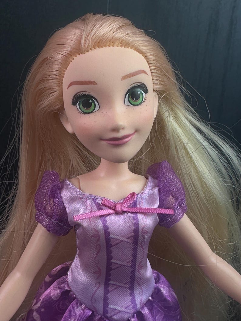 May include: A doll with long blonde hair and green eyes. The doll is wearing a purple dress with a pink bow. The doll has a smiling expression and freckles on its face. The doll's dress has a corset-like design.