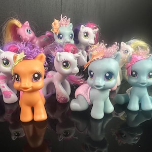Könnte beinhalten: Eine Sammlung bunter Spielzeugponys mit verschiedenen Haarfarben und Accessoires. Die Ponys befinden sich in verschiedenen Posen, einige stehen, andere knien. Die Spielzeuge haben detaillierte Merkmale und sind vor dunklem Hintergrund.
