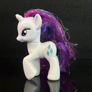 Op de afbeelding: Een wit eenhoornspeelgoed met een paarse en roze manen en staart. Het speelgoed heeft blauwe diamantvormige cutie marks op de flank en een witte hoorn. Het speelgoed staat op een reflecterend zwart oppervlak.