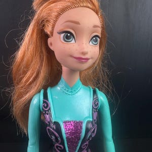 May include: A doll with red hair, freckles, and blue eyes. The doll is wearing a teal and black outfit with a glittery purple center. The doll's hair is styled in a messy updo.