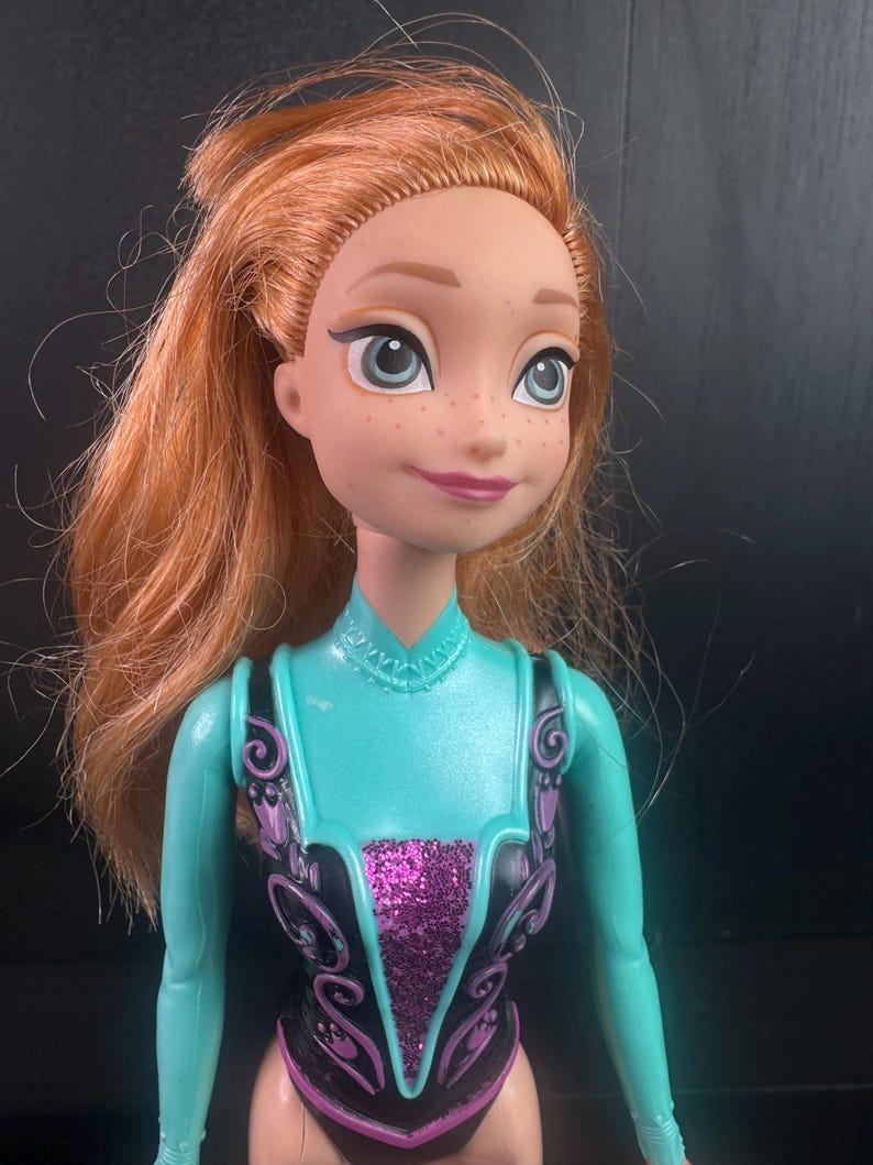 May include: A doll with red hair, freckles, and blue eyes. The doll is wearing a teal and black outfit with a glittery purple center. The doll's hair is styled in a messy updo.