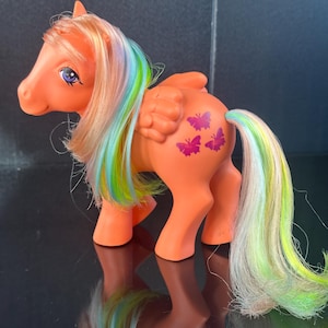 Puede incluir: Poni de juguete naranja con pelo y alas de arcoíris. El poni tiene diseños de mariposas moradas en el flanco. La melena y la cola presentan tonos de amarillo, verde, azul y rosa. Un juguete coleccionable vintage.