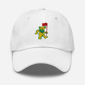 Könnte beinhalten: Weiße Baseballkappe mit einem gestickten gelben Bären, grünem Kragen und roter Flagge. Der Bär wird beim Gehen dargestellt. Die Kappe hat einen gebogenen Schirm und einen verstellbaren Riemen. Das Design ist ein beliebtes Motiv.