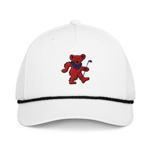 May include: White baseball cap with a black rope detail and a red bear embroidered on the front. The bear is holding a golf club. The bear has a blue collar and the golf club is blue.
