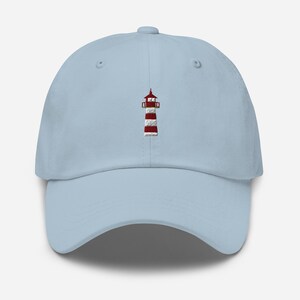 Può includere: Cappellino da baseball azzurro con un ricamo di un faro a strisce rosse e bianche. Il cappellino ha una visiera curva e un cinturino regolabile sul retro. Il design del faro presenta strisce rosse e bianche.