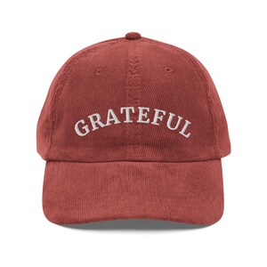 May include: A brick-red corduroy baseball cap with the word "GRATEFUL" embroidered in white across the front. The cap has a curved brim and a rounded top.