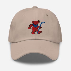 May include: A tan baseball cap featuring an embroidered red bear with a blue collar and hockey stick. The bear is in a dancing pose. The cap has a curved brim and a fabric strap closure.