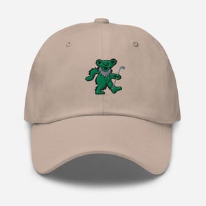 Könnte beinhalten: Beige Baseball Cap mit einem gestickten grünen Bären, der einen Golfschläger hält. Der Bär hat einen weißen Kragen. Die Kappe hat einen gebogenen Schirm und einen verstellbaren Riemen.