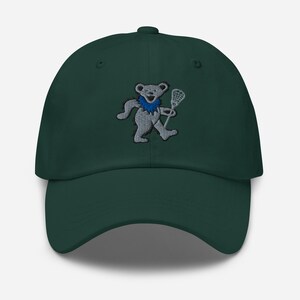 May include: A dark green baseball cap featuring an embroidered bear holding a lacrosse stick. The bear has a blue collar and is in a walking pose. The cap has a curved brim and an adjustable strap.