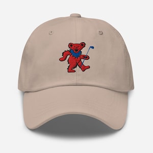 May include: A tan baseball cap featuring an embroidered red bear with a blue collar and golf club. The bear is holding a blue golf club. The cap has a curved brim and a button on top.