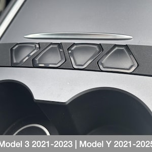 Op de afbeelding: Close-up van het interieur van een Tesla Model 3 of Model Y. De afbeelding toont de bekerhouders en een rij van vijf zwarte knoppen op de middenconsole. De tekst "Model 3 2021-2023 | Model Y 2021-2025" is zichtbaar onderaan de afbeelding.