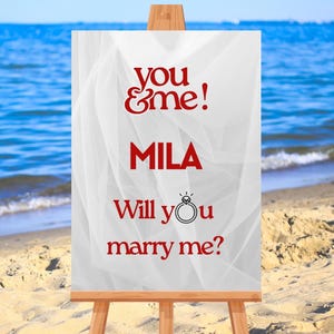 Könnte beinhalten: Eine Holzstaffelei zeigt ein Schild mit den Worten "you & me!" und "MILA" in roter Schrift. Darunter steht auf dem Schild "Will you marry me?" mit einer Ringgrafik. Der Hintergrund ist eine Strandszene.