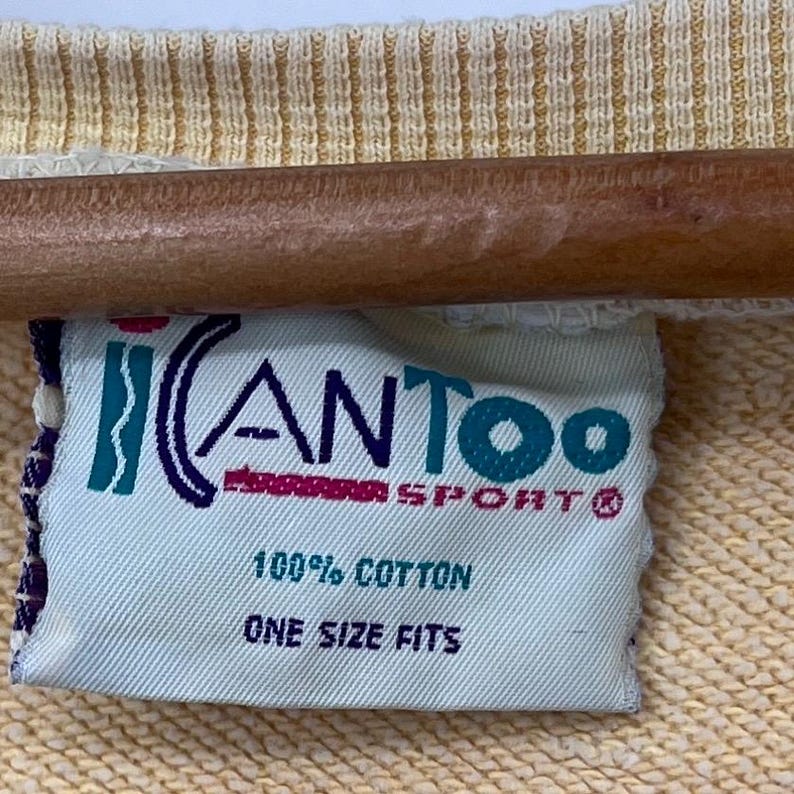 Op de afbeelding: Een wit label met blauwe, roze en turquoise letters met de tekst "iCANTOO SPORT&reg;" en "100% KATOEN &Eacute;&Eacute;N MAAT".