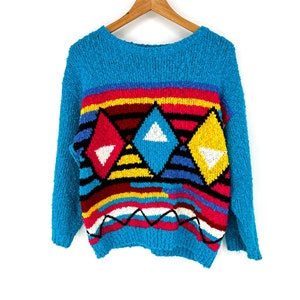 May include: A turquoise sweater with a crew neck and long sleeves. The sweater features a geometric design with red, yellow, blue, and black diamond shapes and horizontal stripes. The sweater is hanging on a wooden hanger.