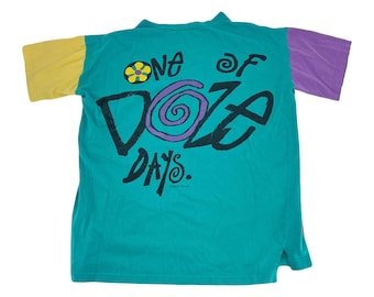 Könnte beinhalten: Ein türkisfarbenes T-Shirt mit gelben und lila Ärmeln. Auf der Rückseite des T-Shirts steht in Schwarz der Text "ONE OF DOZE DAYS", mit einem lila Wirbeldesign und einer gelben Blumengrafik. Das Shirt ist im Vintage-Stil.