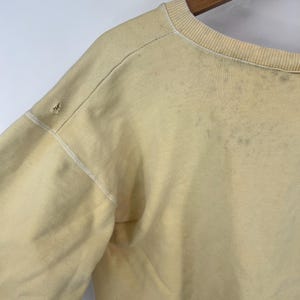 Op de afbeelding: Een lichtgeel sweatshirt met een ronde hals en lange mouwen. Het sweatshirt heeft een verbleekte uitstraling en enkele zichtbare vlekken.