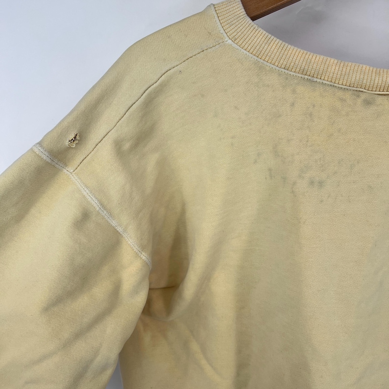 Op de afbeelding: Een lichtgeel sweatshirt met een ronde hals en lange mouwen. Het sweatshirt heeft een verbleekte uitstraling en enkele zichtbare vlekken.