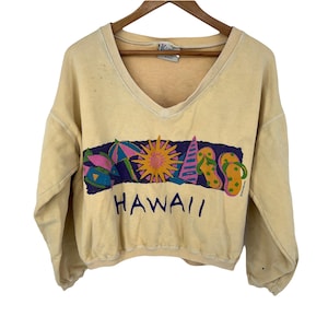 Op de afbeelding: Een lichtgeel, gecropped sweatshirt met een v-hals en een kleurrijke grafische print van een zon, parasols, vissen en slippers met het woord "HAWAII" eronder gedrukt.
