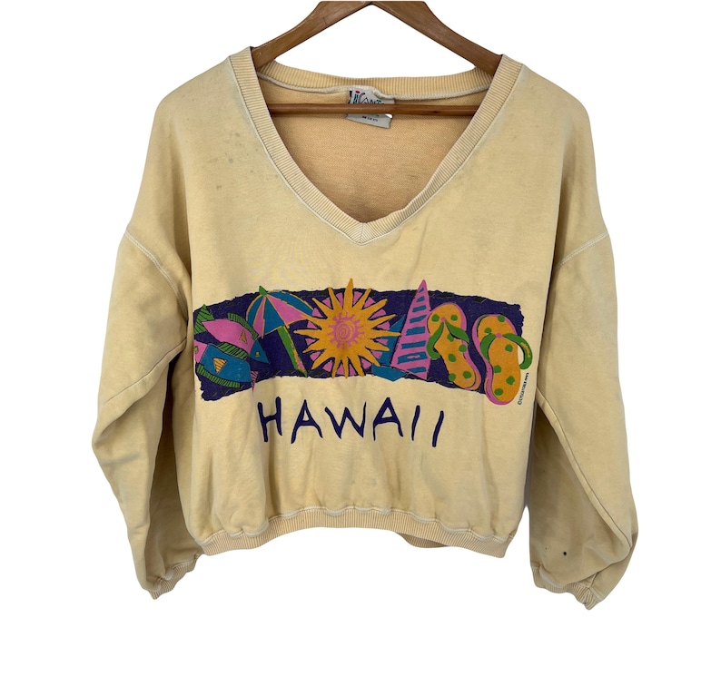 Op de afbeelding: Een lichtgeel, gecropped sweatshirt met een v-hals en een kleurrijke grafische print van een zon, parasols, vissen en slippers met het woord "HAWAII" eronder gedrukt.