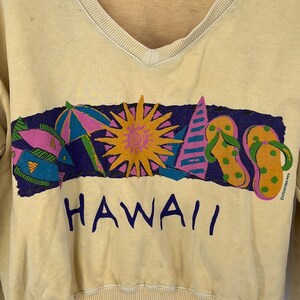 Op de afbeelding: Een lichtgeel sweatshirt met een kleurrijke grafische print van een zon, een vis, een paraplu en slippers. De grafiek is omgeven door een blauwe rand en het woord "HAWAII" is eronder gedrukt.