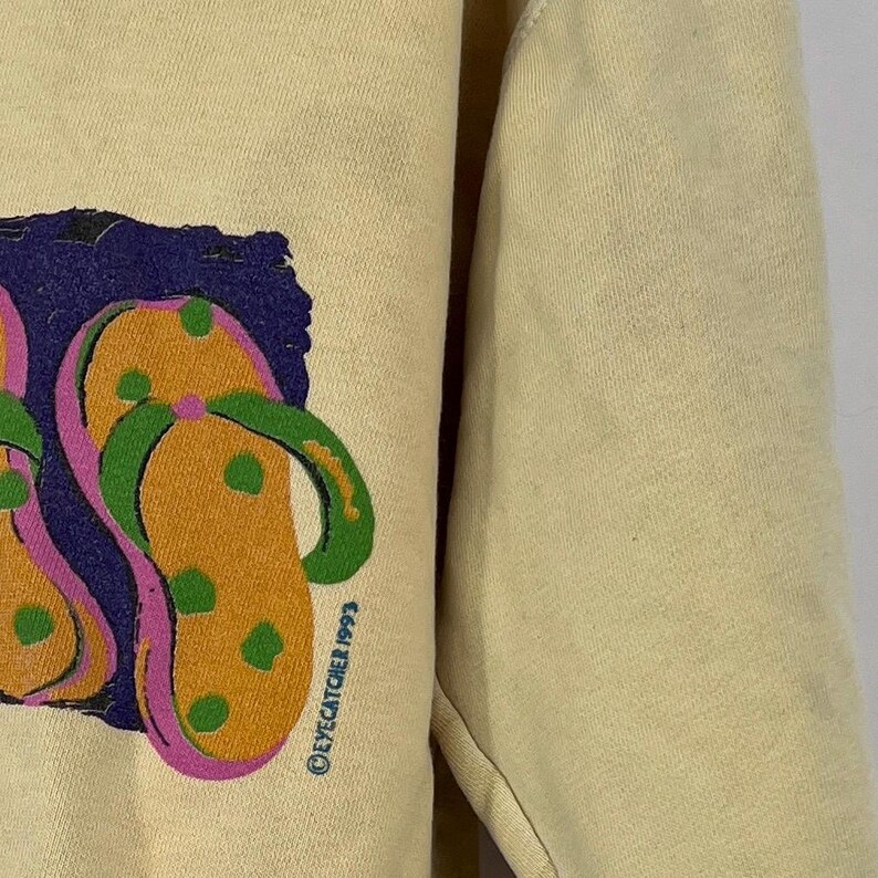 Op de afbeelding: Een geel sweatshirt met een afbeelding van twee kleurrijke slippers. De afbeelding bevat de tekst "Eyecatcher 1993".