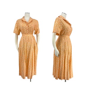 Op de afbeelding: Een vintage oranje jurk met een witte bloemenprint en een knoopsluiting aan de voorkant. De jurk heeft korte mouwen en een getailleerde pasvorm met een riem.