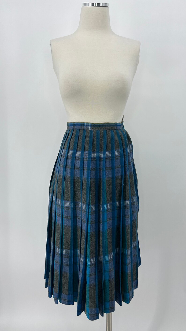 May include: A knee-length pleated skirt in a blue, gray, and teal plaid pattern. The skirt is displayed on a mannequin against a white background. The fabric has a textured appearance.