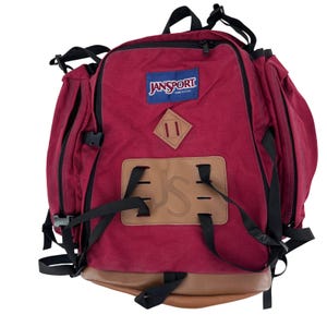 May include: A burgundy Jansport backpack with black straps and accents. The backpack features a brown leather bottom and a brown leather patch with the letters "JS" embossed. The Jansport logo is on a blue rectangle.