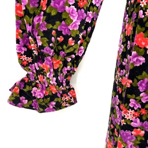 May include: Close-up of a black floral print garment. The fabric features a pattern of purple, red, and green flowers. The sleeve has a ruffled cuff. The garment appears to be made of a soft, velvety material.