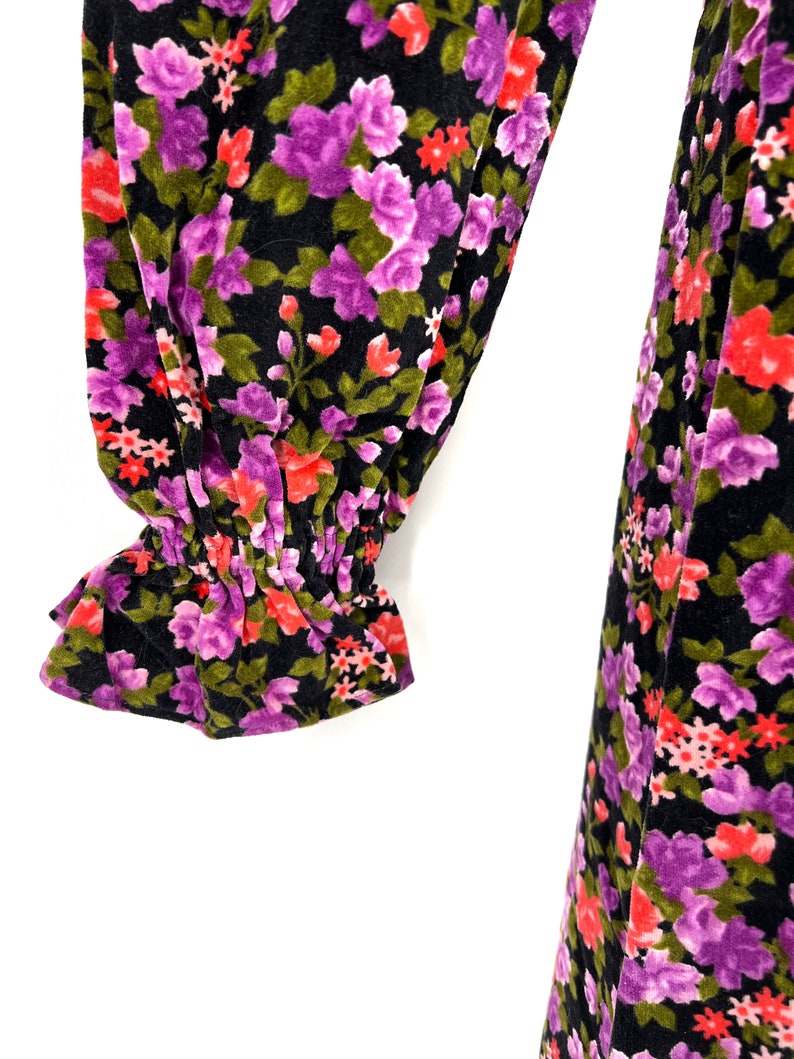 May include: Close-up of a black floral print garment. The fabric features a pattern of purple, red, and green flowers. The sleeve has a ruffled cuff. The garment appears to be made of a soft, velvety material.