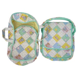 May include: A quilted baby carrier and matching diaper bag set. The set features a pastel color palette with a repeating pattern of baby dolls, balloons, and toys. The carrier has a handle and a soft interior.