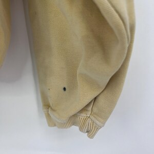 Op de afbeelding: Close-up van de onderste zoom van een lichtbeige sweatshirt. De stof is zacht en heeft een licht verweerde look. Er zijn een paar kleine donkere vlekken op de stof.