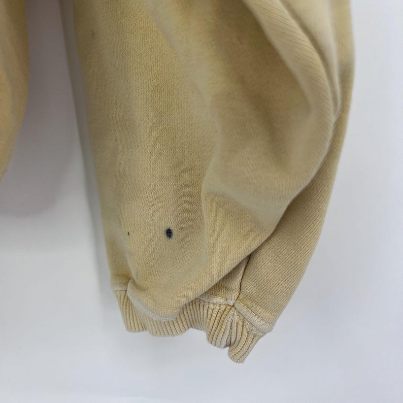Op de afbeelding: Close-up van de onderste zoom van een lichtbeige sweatshirt. De stof is zacht en heeft een licht verweerde look. Er zijn een paar kleine donkere vlekken op de stof.