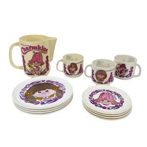 Op de afbeelding: Een vintage kindertheeset met een kan, kopjes en borden. De set is crèmekleurig met illustraties van een meisje met een roze hoed en het woord "Charmkins". De borden en kopjes hebben een bloemenrand.