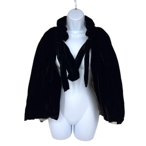 May include: A black velvet cape with a rounded collar and long ties. The cape is displayed on a white mannequin, showcasing its flowing design. The fabric appears soft and luxurious, suitable for formal wear or costume.
