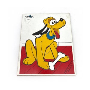 May include: A vintage Playskool Pluto puzzle featuring a yellow cartoon dog with a blue collar and a bone. The puzzle has a white background with a red border and the text "Playskool" and "Pluto". Made in the U.S.A.