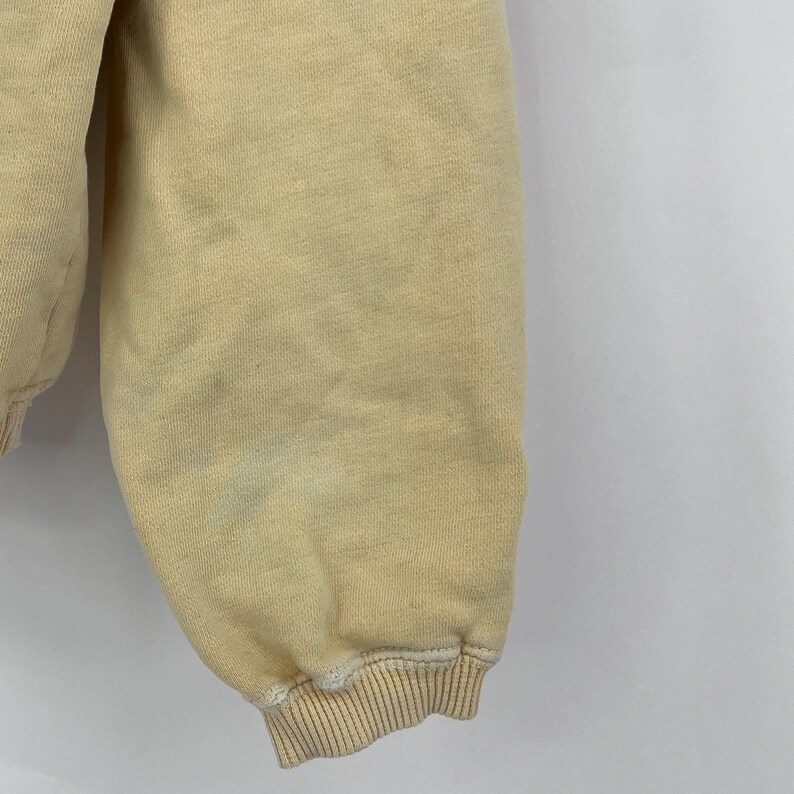 Op de afbeelding: Close-up van de boord van een lichtgeel sweatshirt. De stof is zacht en licht getextureerd. De boord is omgevouwen en heeft een geribbelde textuur.